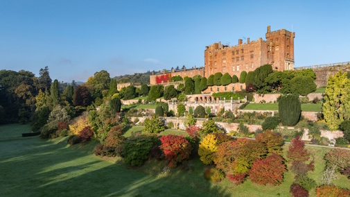 Wide view of Powis Castle rising above its terraced gardens in autumn. Manicured yew topiary and stone terraces cascade down the hillside, while trees and shrubs in shades of red, orange, gold and green fill the foreground. Early morning sunlight illuminates the castle and garden against a clear blue sky.